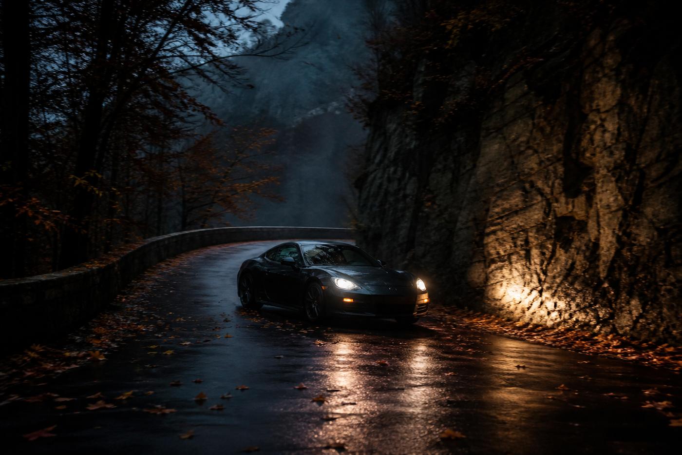 Dark sports car on a wet hairpin, headlights raking a rock wall at dusk.