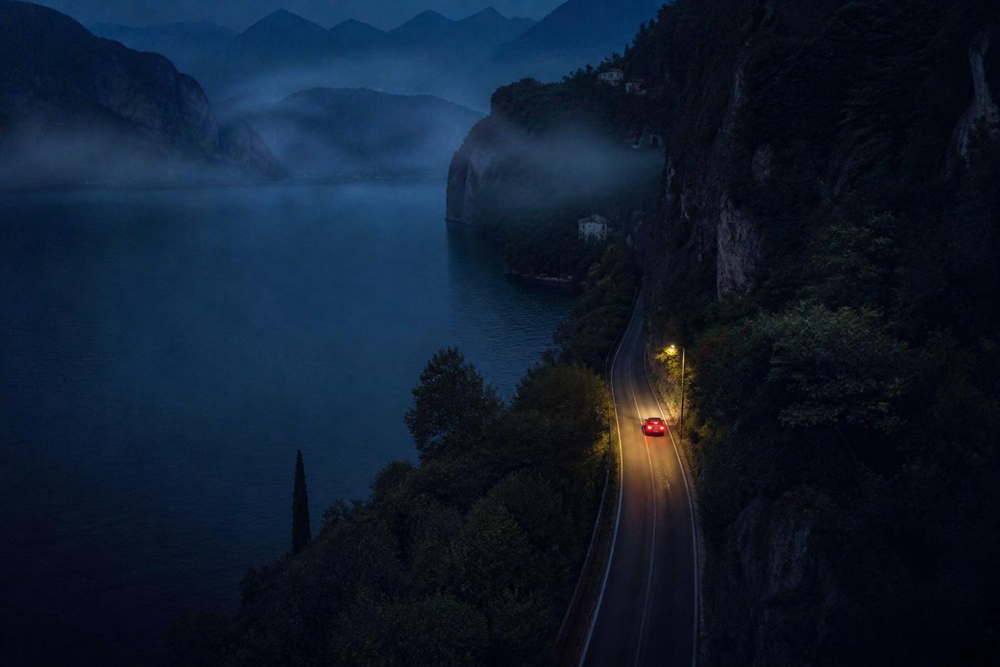 High view of a canyon road hugging a dark lake, taillights under a single streetlamp.