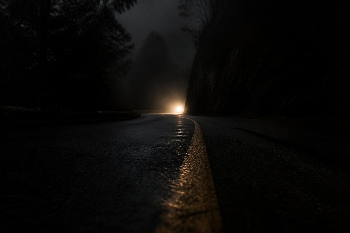 Low view down a yellow center line toward a headlight bloom around a canyon bend.